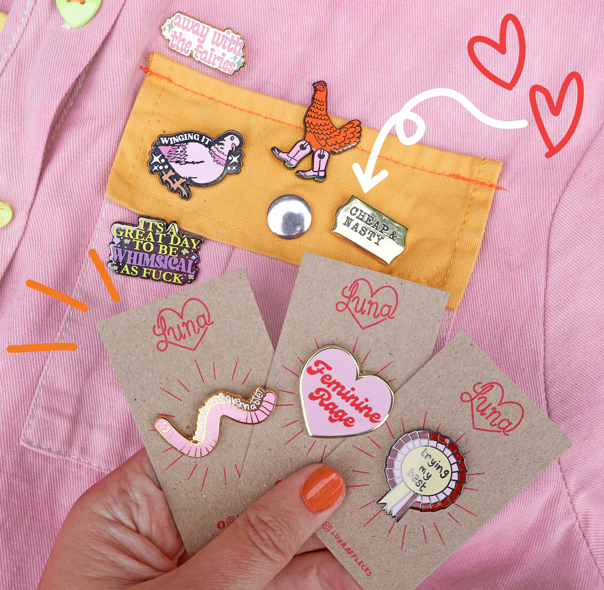 A hand holds three brown display cards showing colorful novelty enamel pins, with a matching yellow card above showing more pins pinned to a pink jacket. The pins feature playful slogans and illustrations including a pink worm, a heart-shaped 'Feminine Rage' pin, and a circular pin with a clock face.